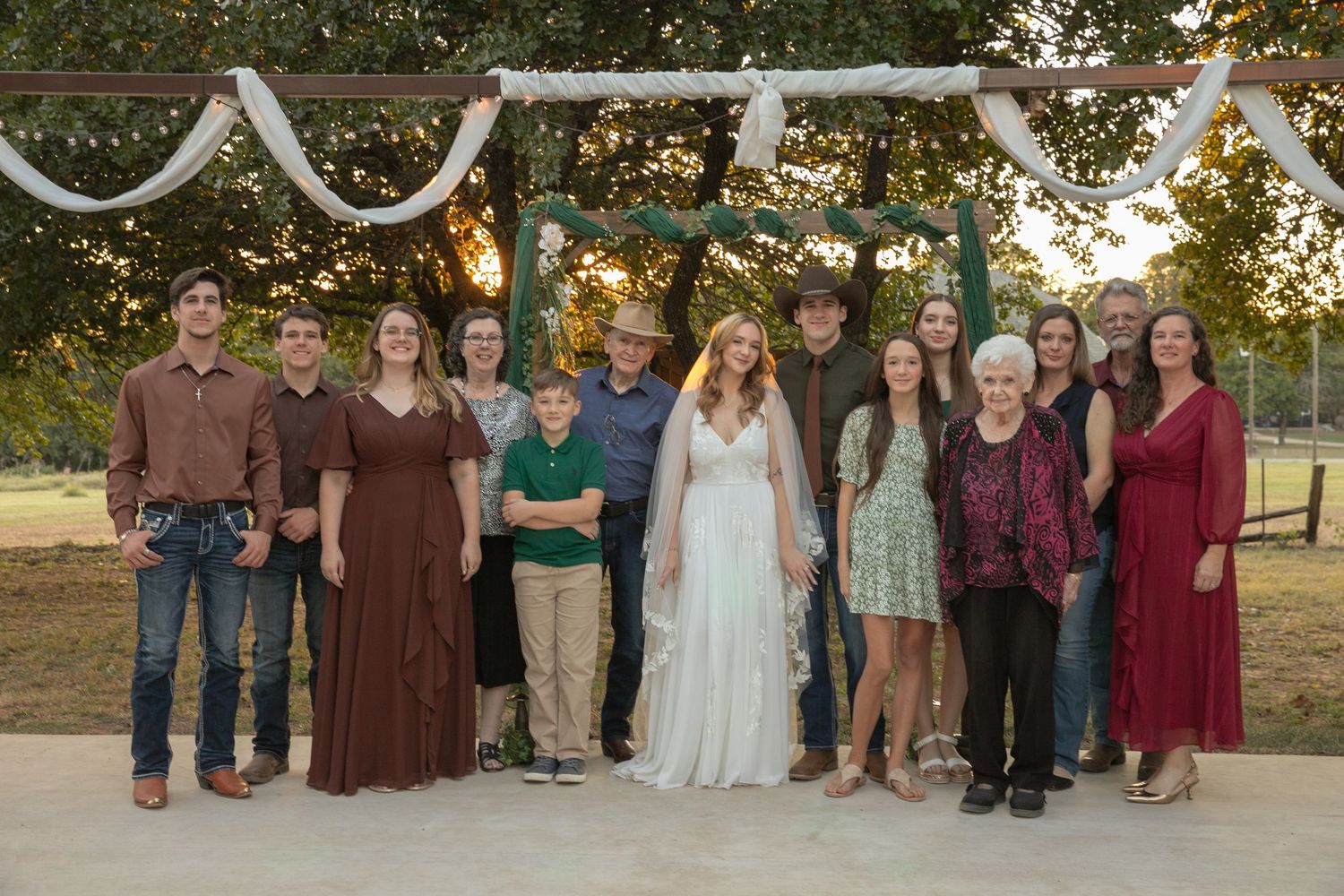 A large wedding party group photo under a rustic floral arch at an outdoor sunset wedding celebration.