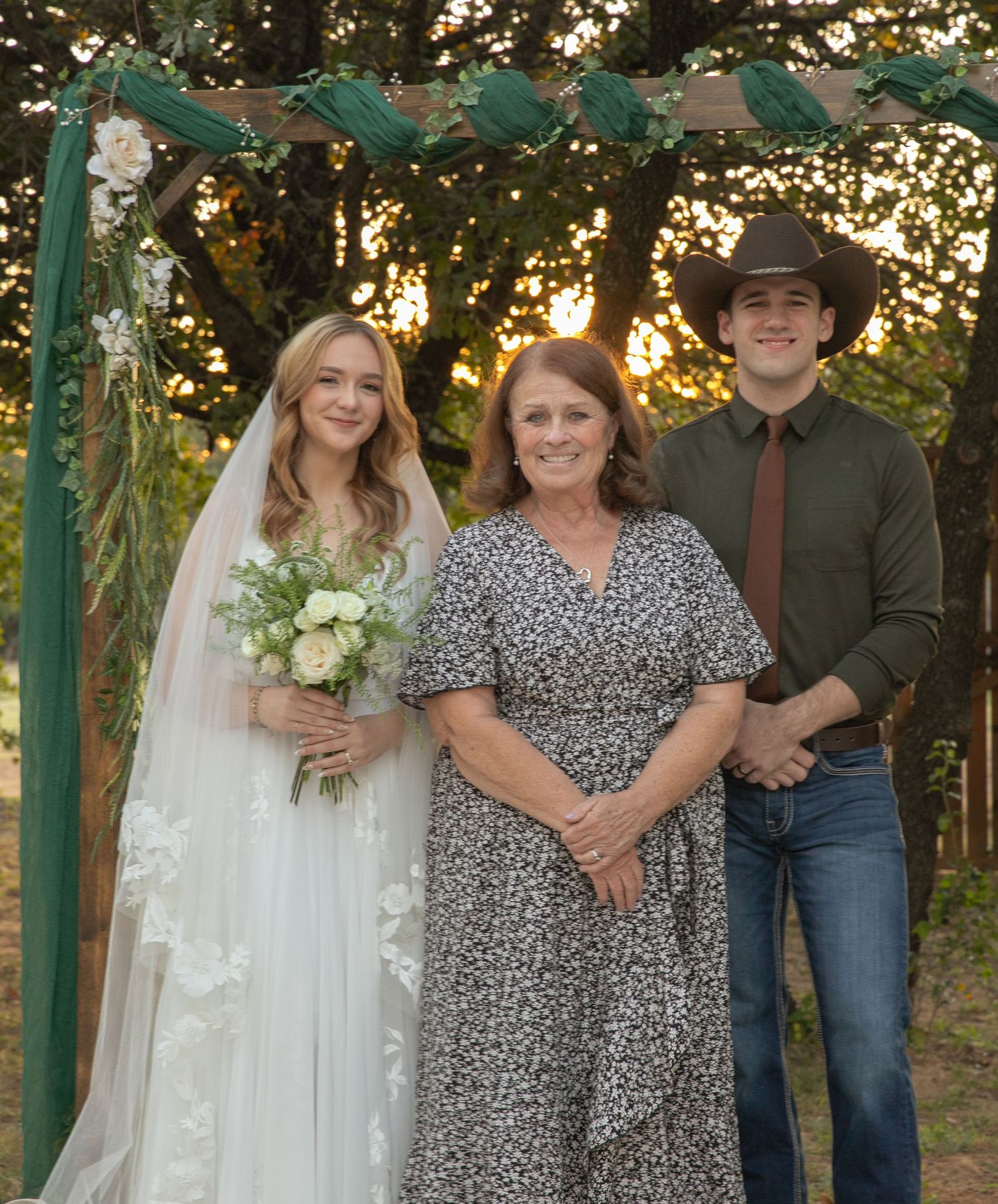 A blonde bride in a white gown and veil poses with a woman in floral dress and young man under a floral arch.