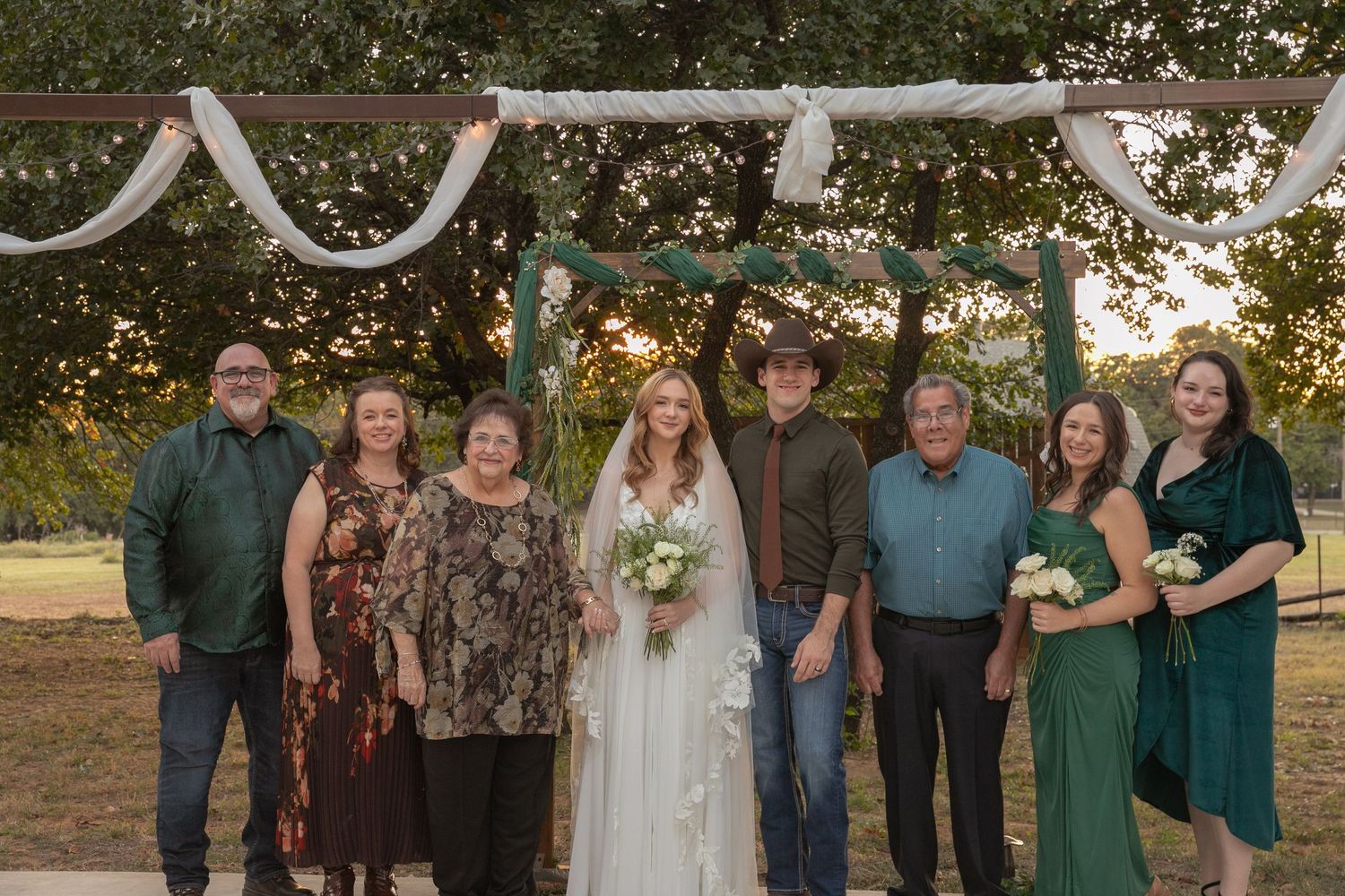 A bride and groom in a cowboy hat pose with family members under a rustic arch at a golden hour wedding.