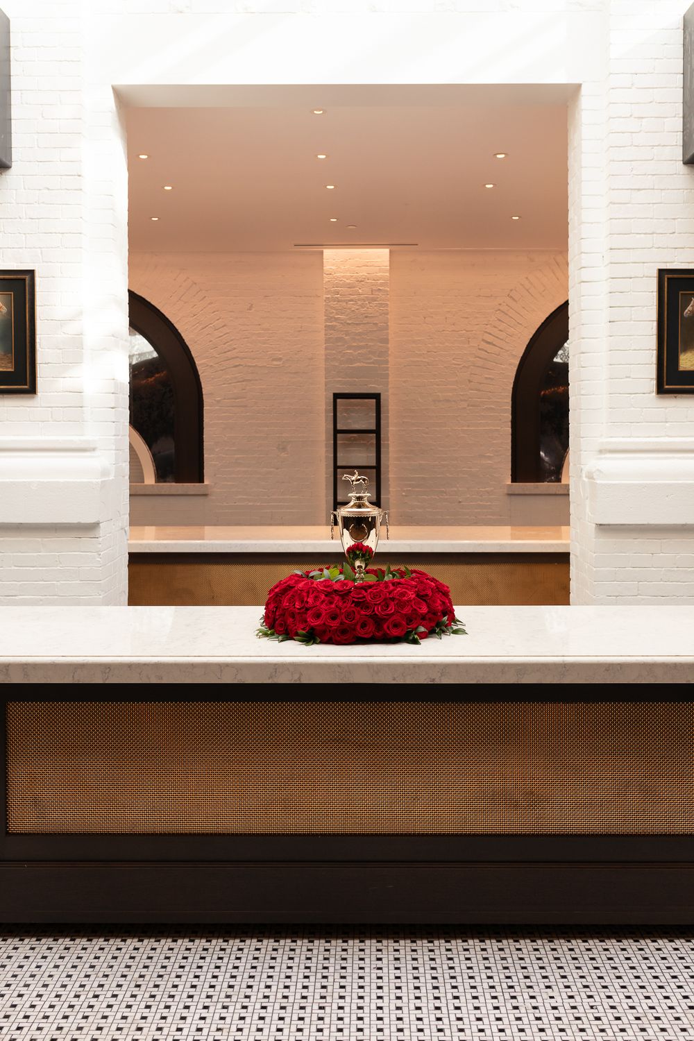 Elegant hotel lobby with red roses arrangement and silver trophy on marble counter, white brick walls with arched details.