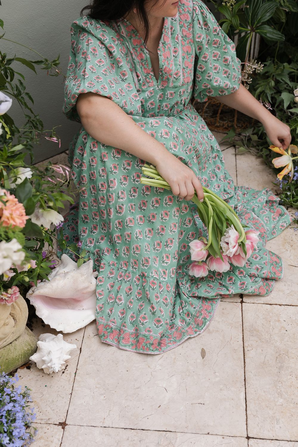 Someone in a vintage-style mint green floral dress arranges fresh pink tulips while sitting on stone pavers outdoors.