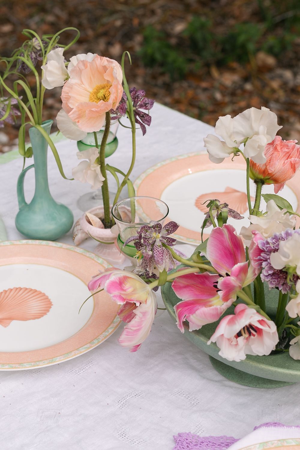 Elegant table setting with peach and pink floral arrangements in turquoise vases alongside seashell-patterned plates on white linen.