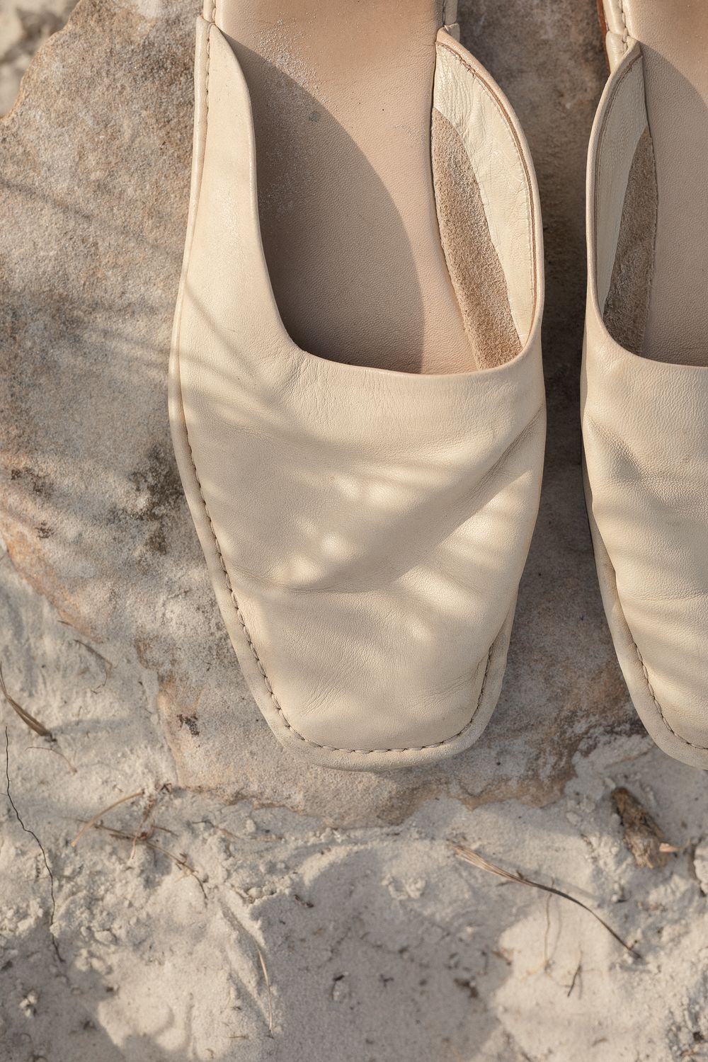 Close-up view of cream colored leather slip-on shoes with square toes resting on a textured stone surface.
