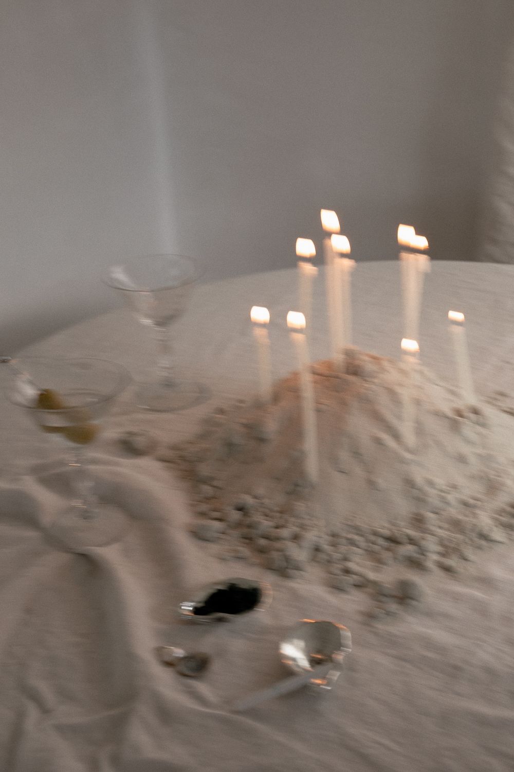 Ethereal table scene with lit taper candles, sand, crystals, and a glass on white linen in soft candlelight.