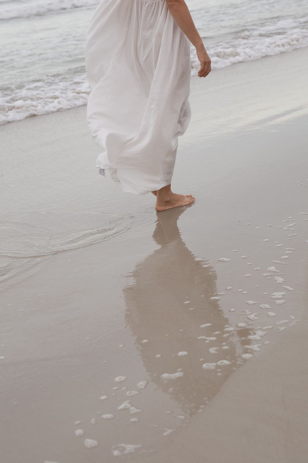 Woman in white dress walking barefoot on wet sandy beach with ocean waves in background.