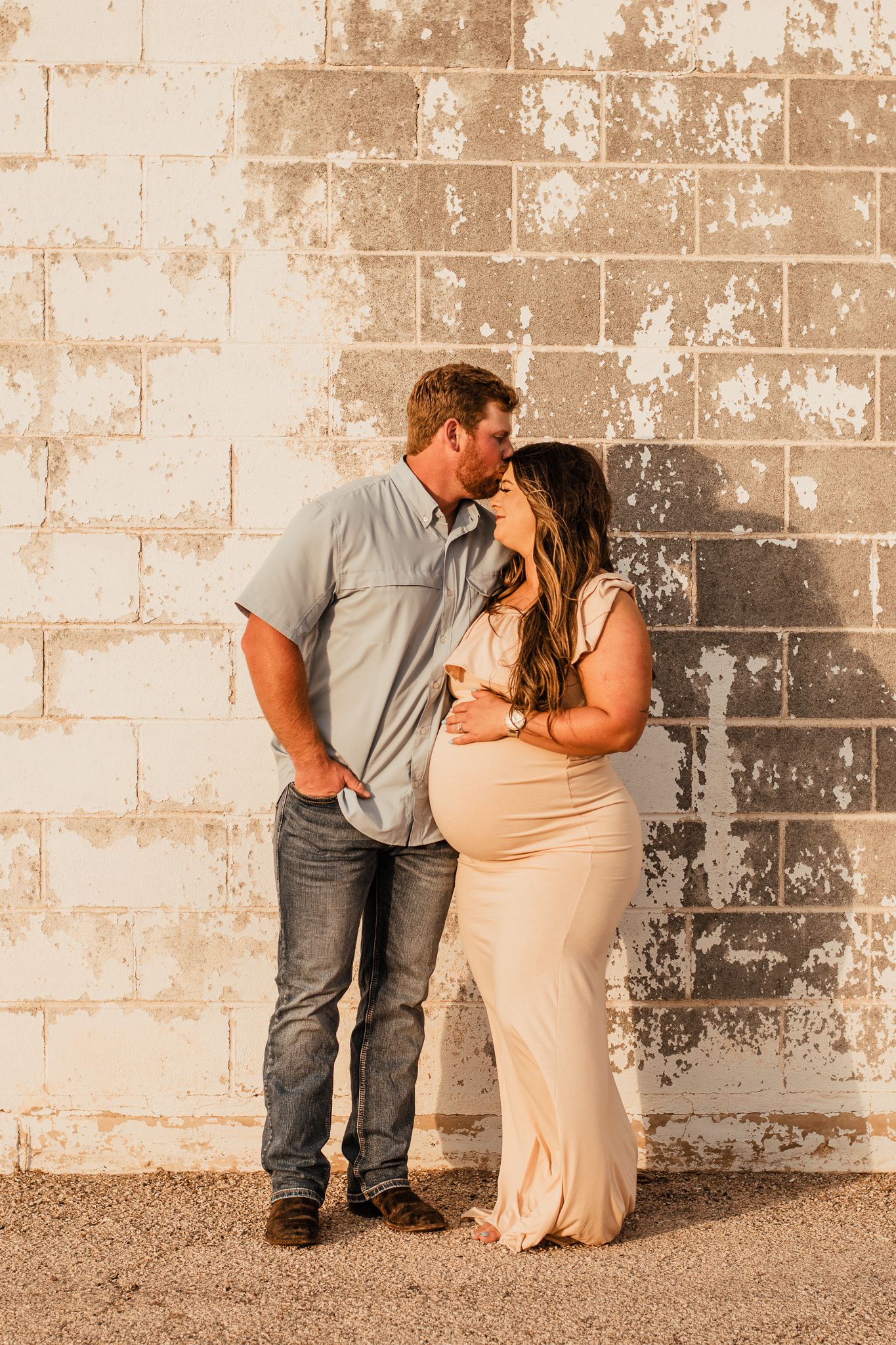 A romantic maternity photoshoot of a couple embracing against a weathered brick wall at sunset.