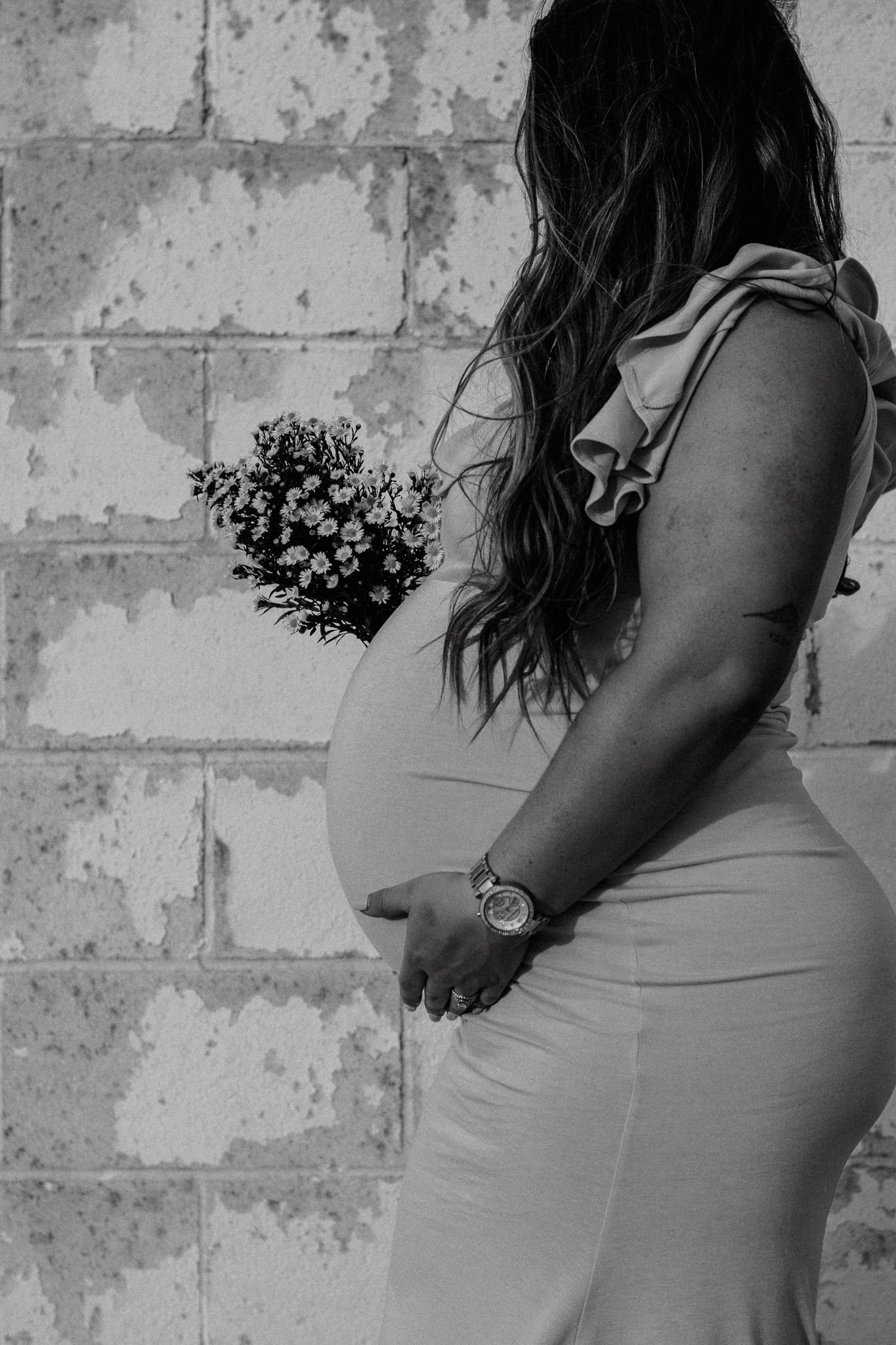 Black and white maternity photo of pregnant woman holding flowers against textured wall.
