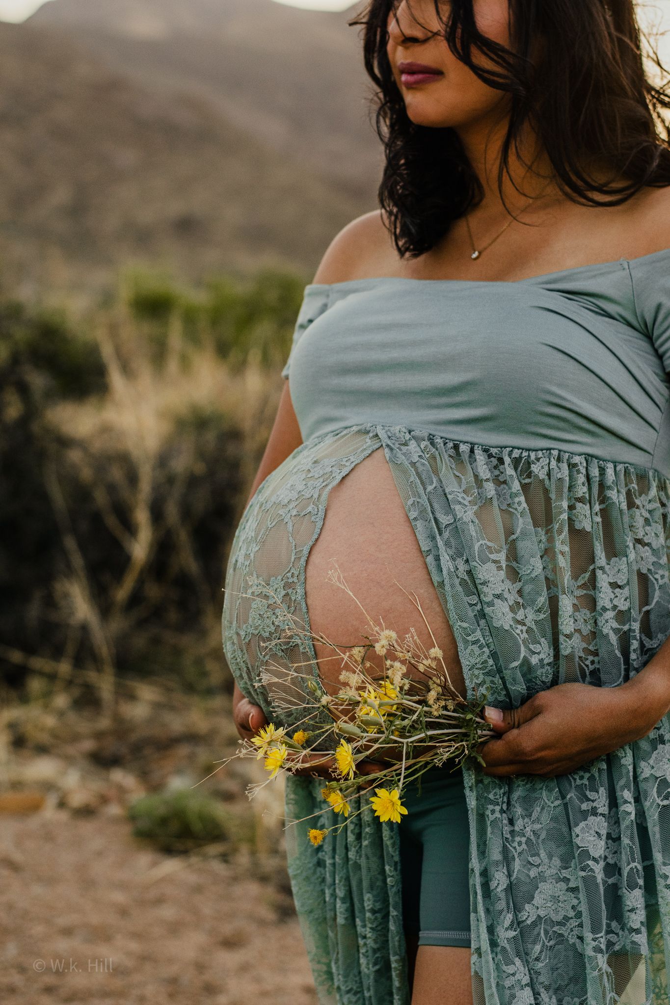 A pregnant woman in a mint green lace dress holds yellow wildflowers against her belly.