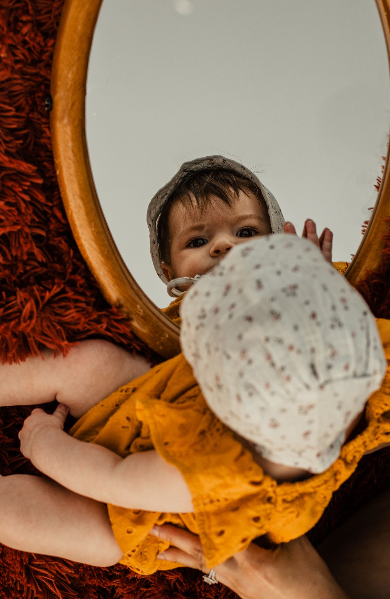 A reflection in a round mirror shows a baby in orange clothing and a bonnet.