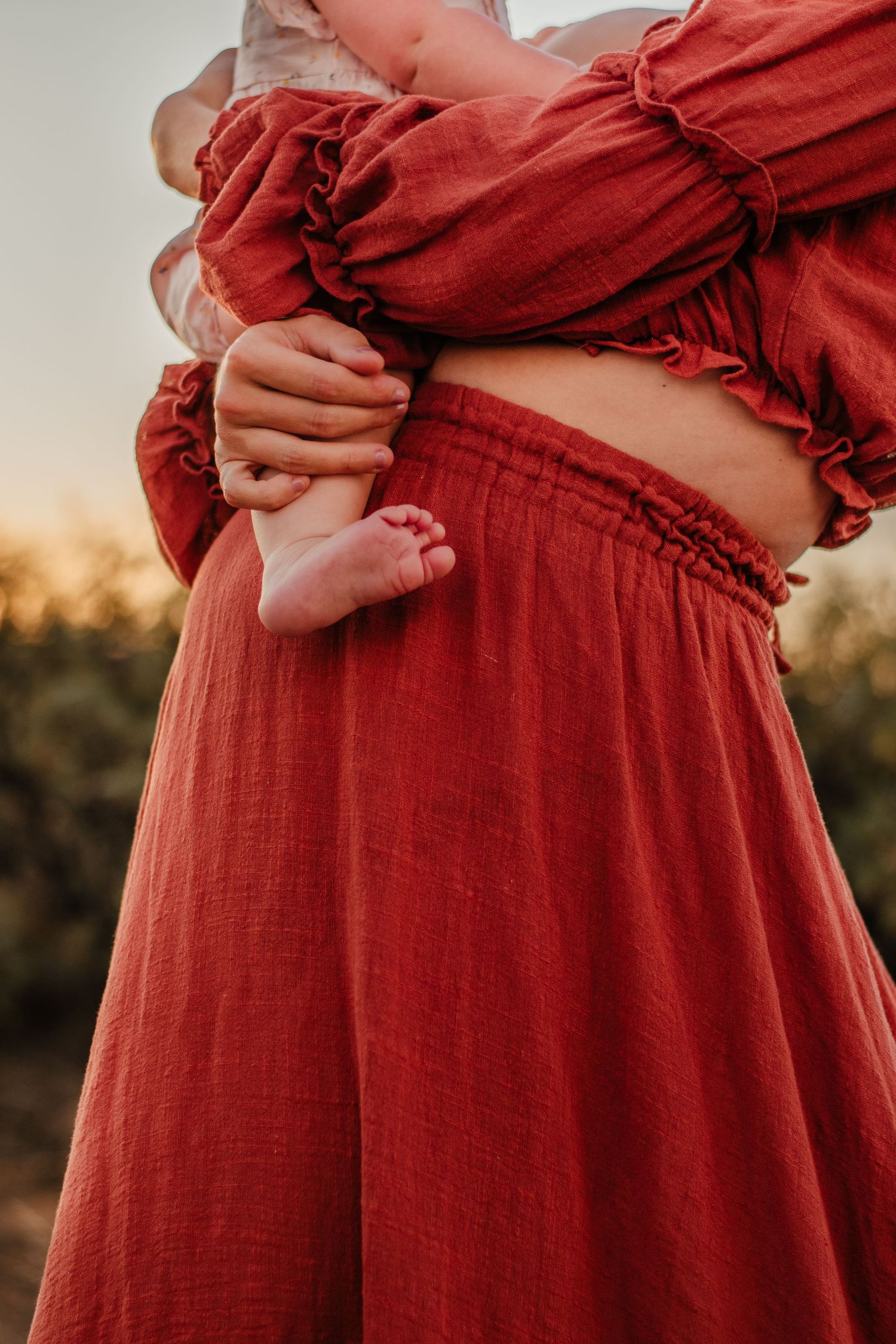 Close-up of maternity photo showing a red flowing dress and baby feet against sunset.