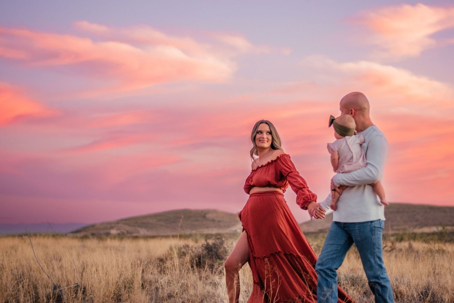 A couple and young child standing together in a field during a vibrant pink sunset.
