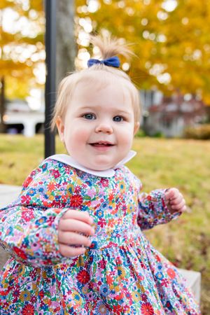 Baby wearing colorful floral dress smiling outdoors during autumn.