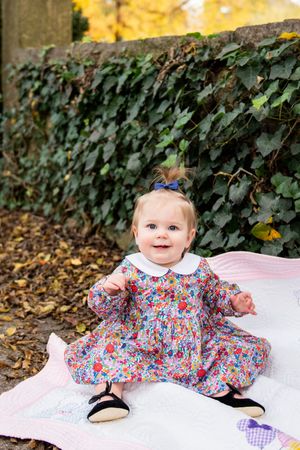 Baby in floral dress lying on pink blanket with ivy covered wall and fall foliage in background.