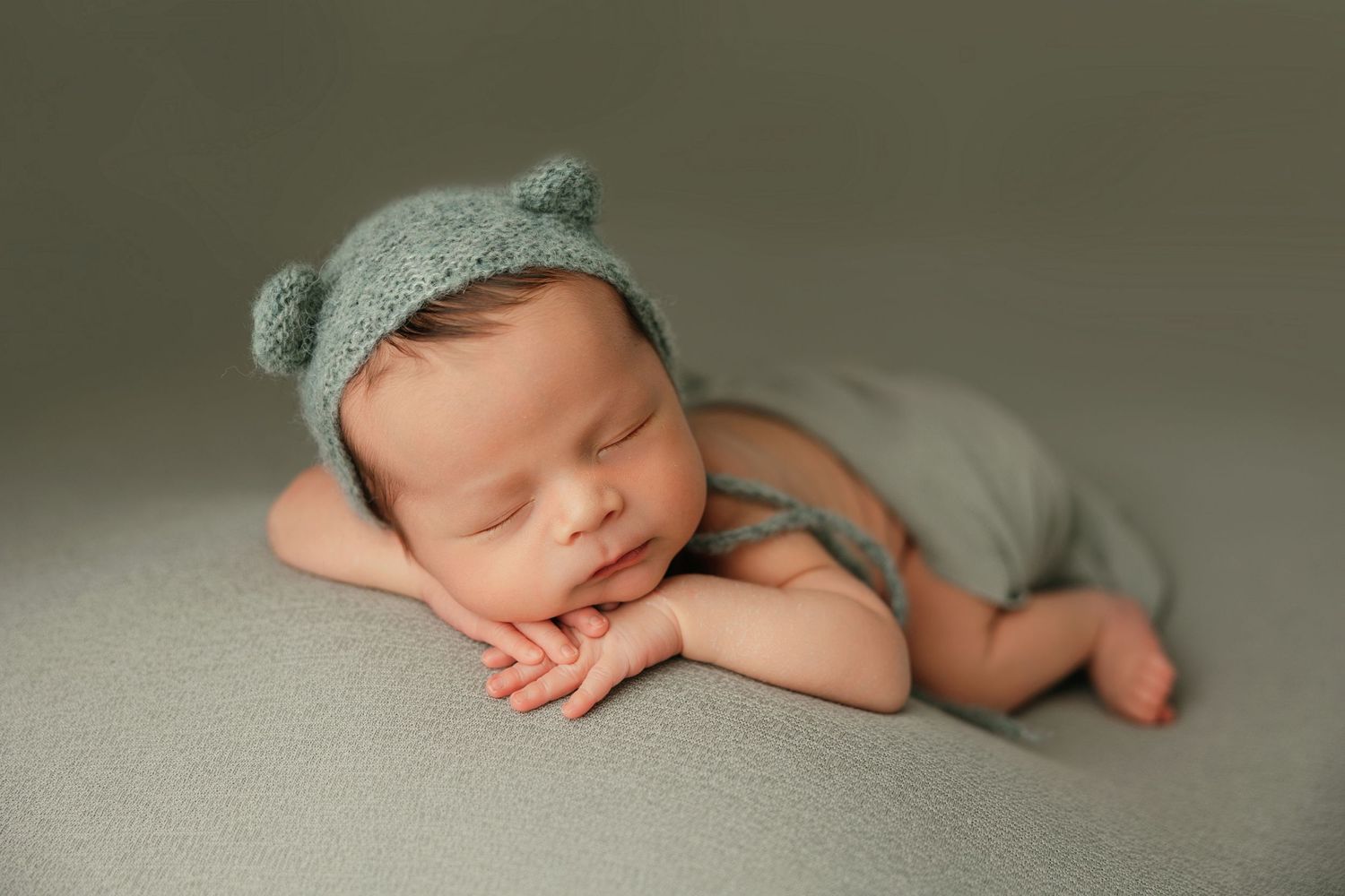 Sleeping newborn in sage green outfit and hat lying on soft gray backdrop.