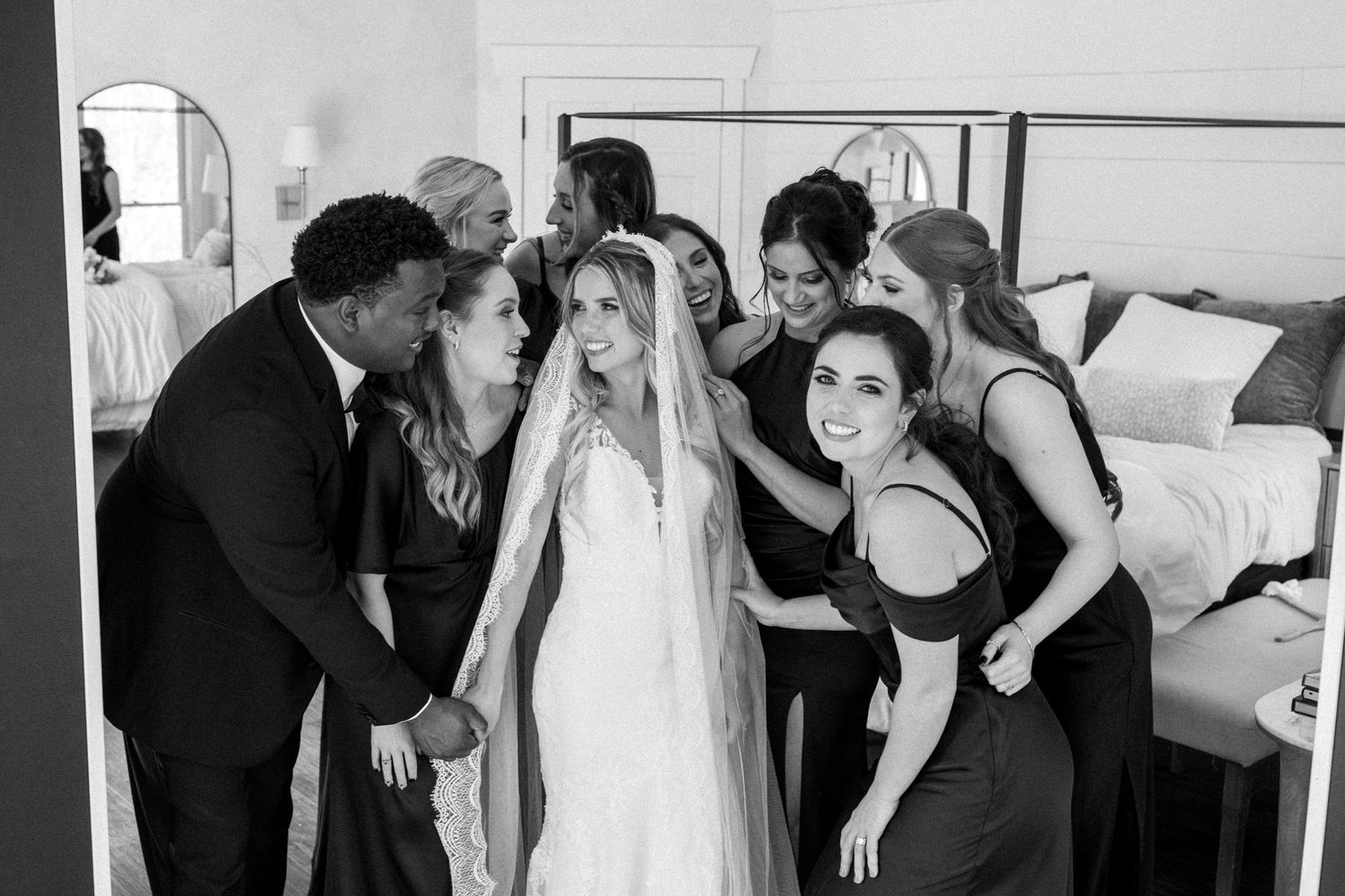 Wedding party gathers around bride in white dress sharing an intimate candid moment before the ceremony.