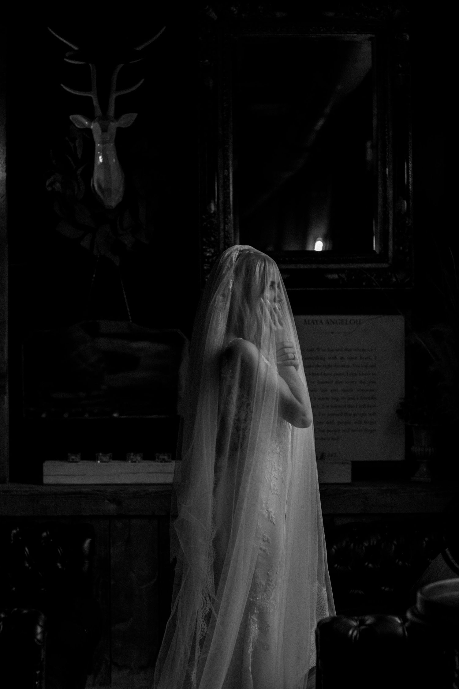 Dramatic black and white photo of bride in flowing veil standing before mounted deer head.