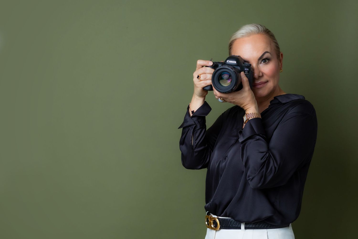 A photographer holds up a Canon DSLR camera to take a photo against a neutral beige background.