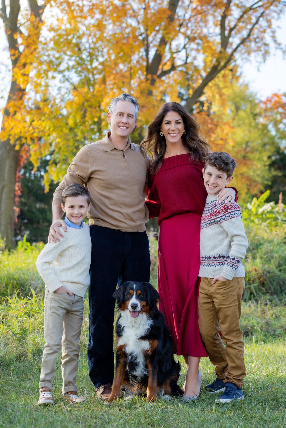 A family poses outdoors with their Bernese Mountain Dog against a backdrop of autumn trees with golden foliage.