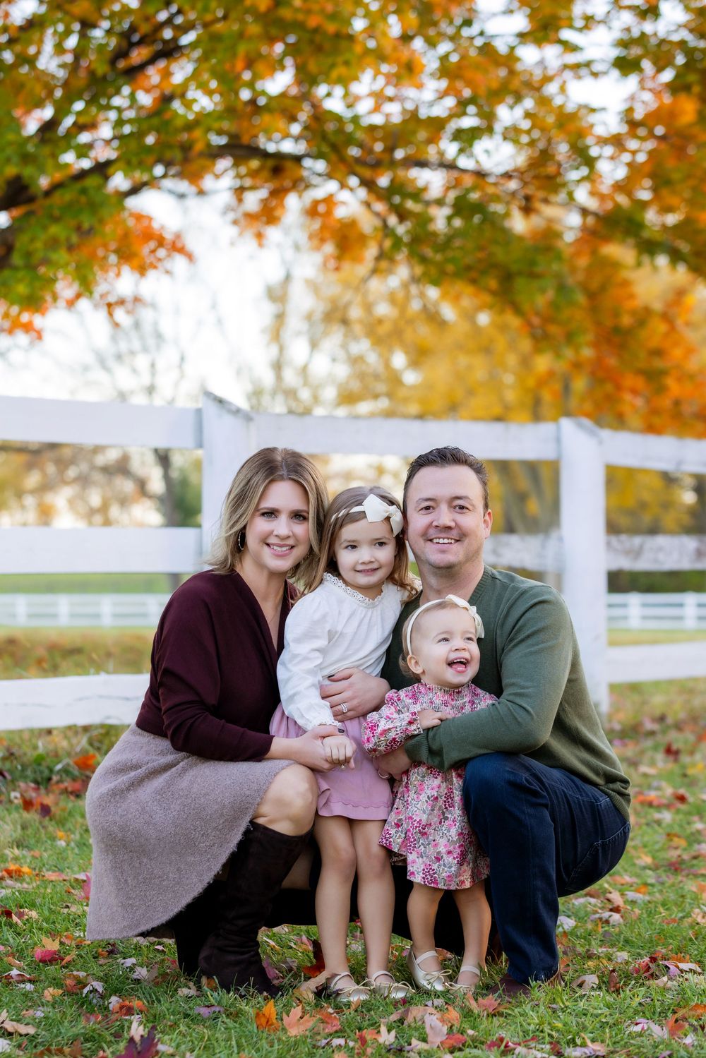 A family sits together on grass surrounded by autumn leaves and white fencing on a fall day.