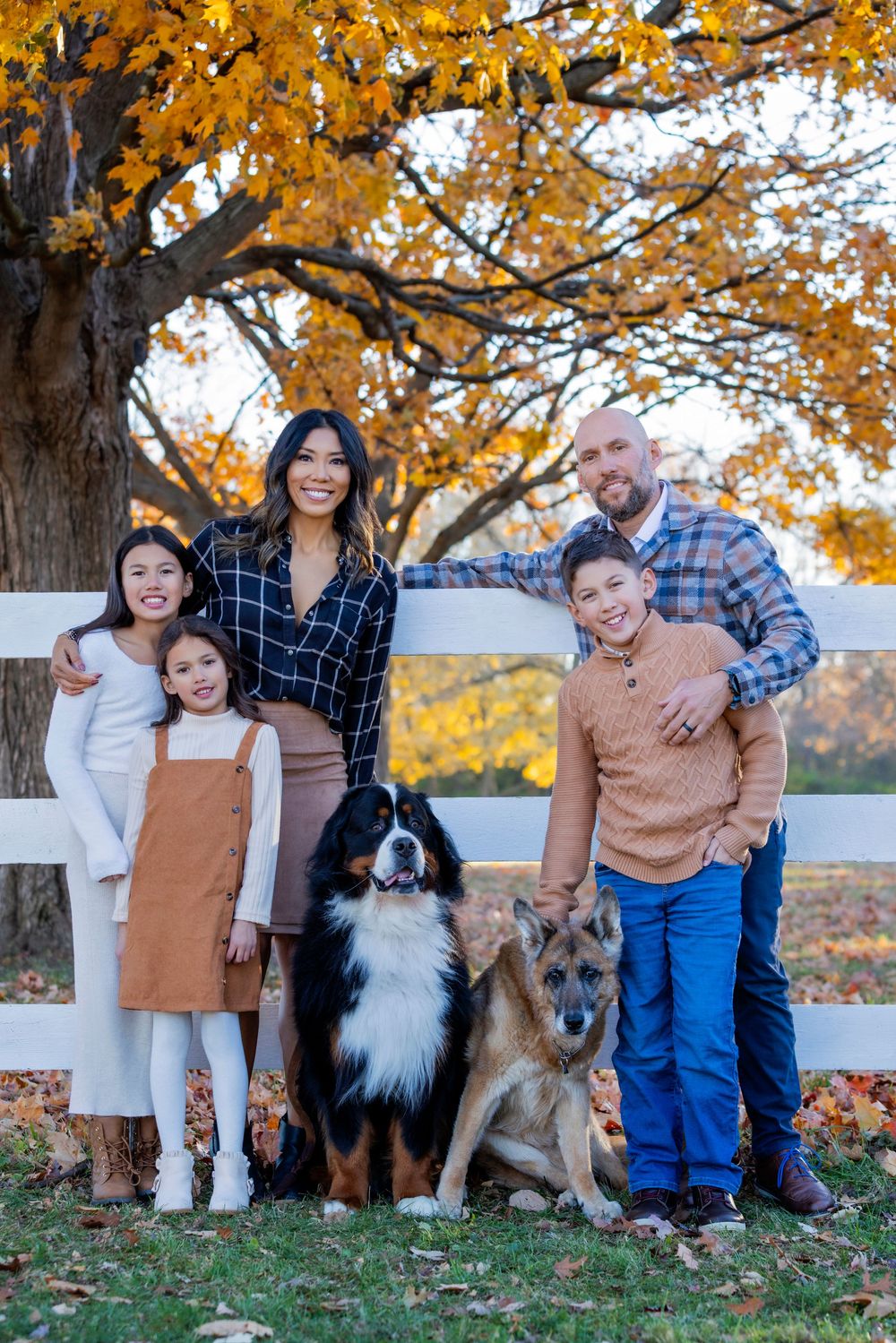 Family and pets pose together on autumn day with golden leaves and white fence in background.