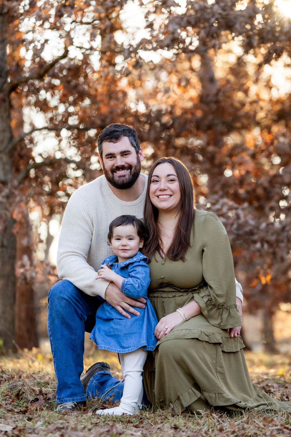 A family sits together on autumn ground with golden sunlight filtering through trees in the background.