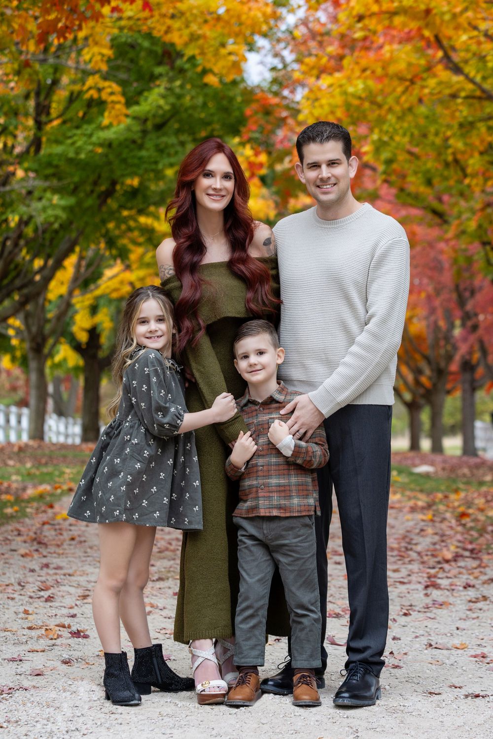 A family stands together on fallen autumn leaves with vibrant orange and red foliage in the background.