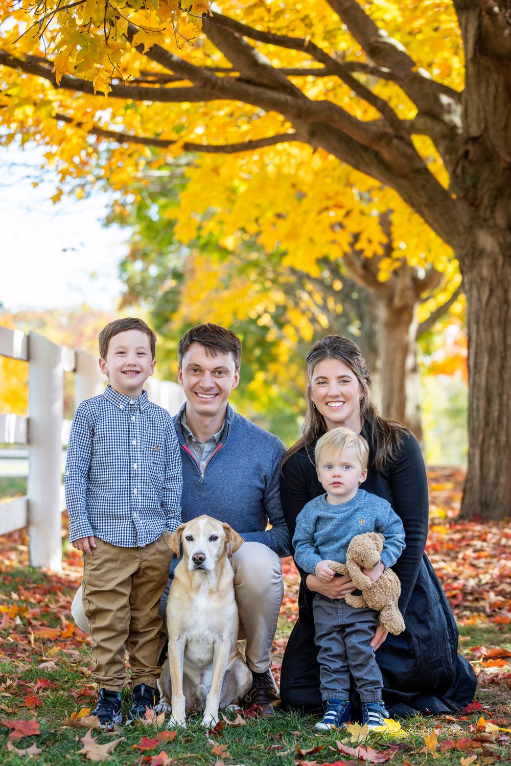 Family sits together under vibrant yellow autumn trees with their white dog and teddy bear on fallen leaves.