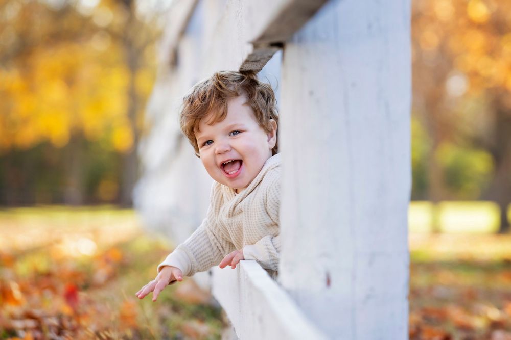 Joyful toddler peeks around white column outdoors during autumn with golden foliage in background.
