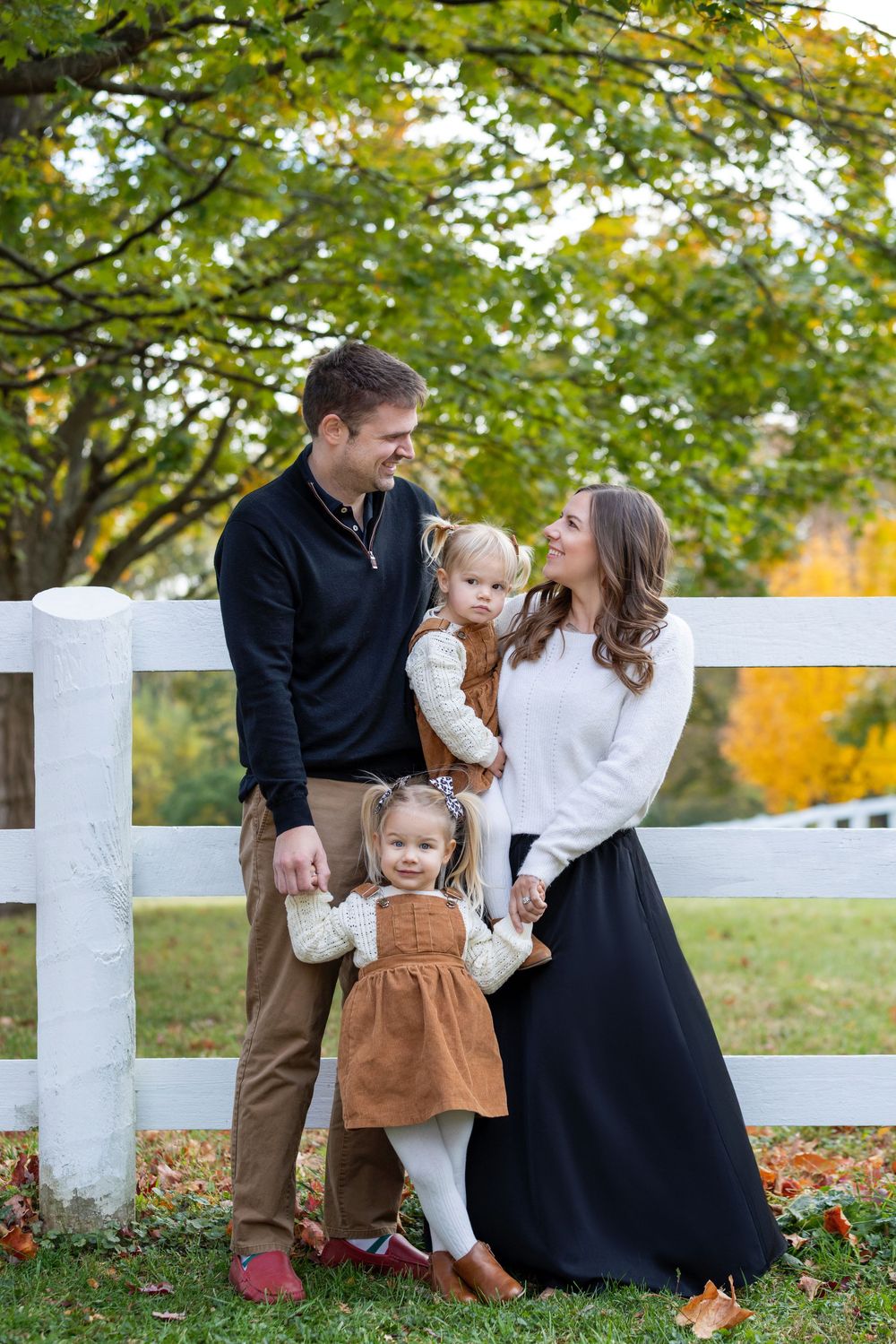 Family in coordinated fall outfits poses together by white fence during autumn photo session in park.