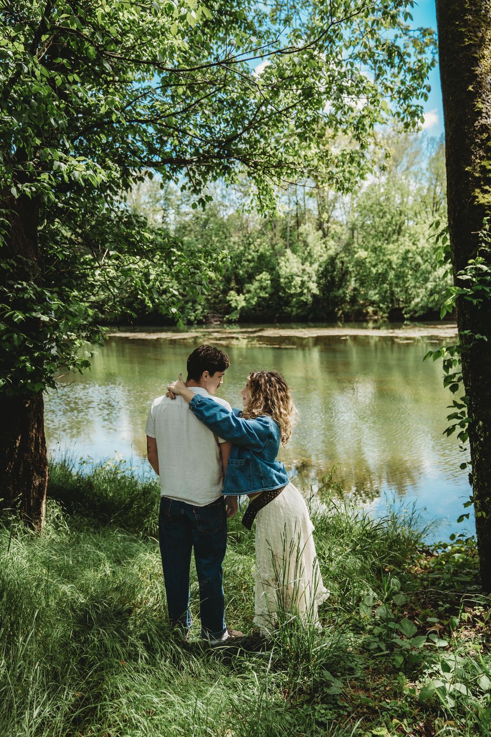 Couple embracing by a tranquil forest lake surrounded by lush green trees on a sunny day.