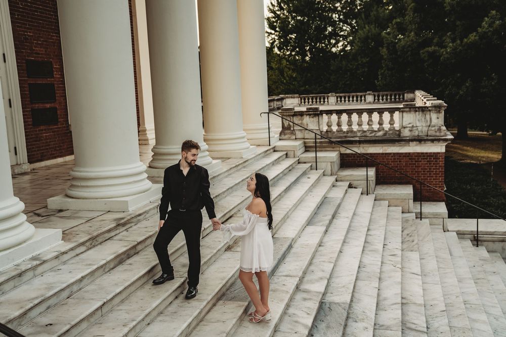 Couple holding hands on grand marble steps with white columns during romantic outdoor engagement photo session.