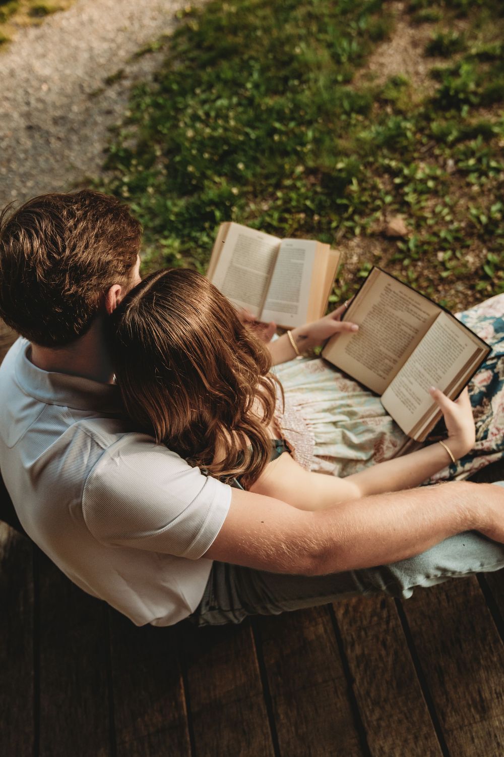 A couple sits on wooden steps outdoors, leaning together while reading open books in a lush green garden setting.