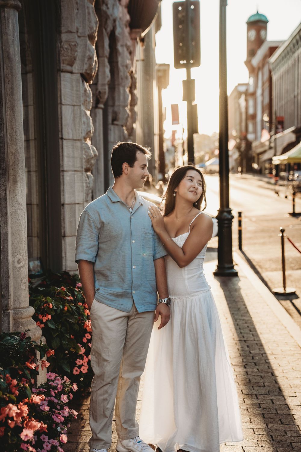 A romantic couple in wedding attire walks along a sunlit cobblestone street with ornate lamp posts and blooming flowers.