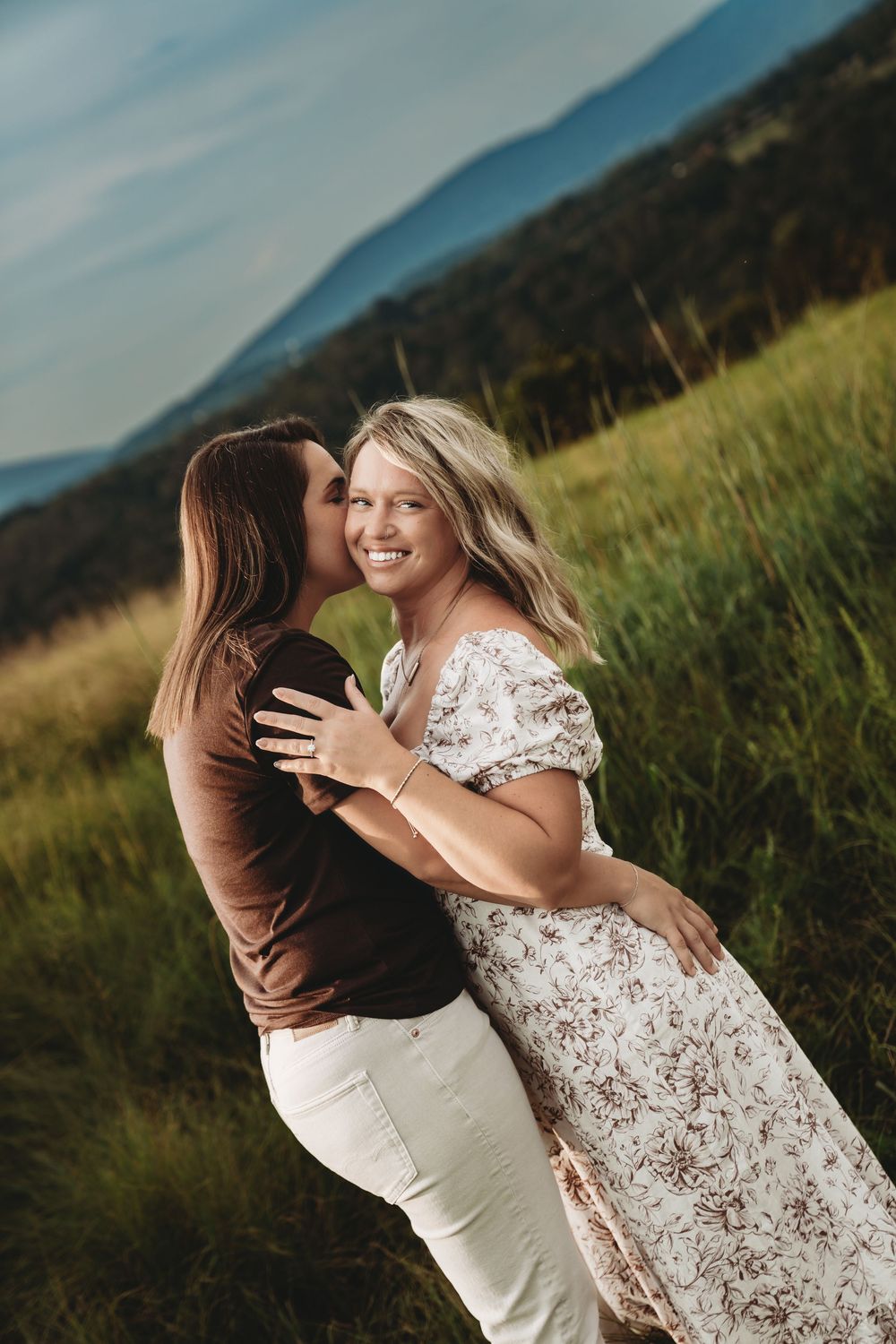 A couple embraces in a sunlit meadow with mountains in the background, wearing brown and white outfits.