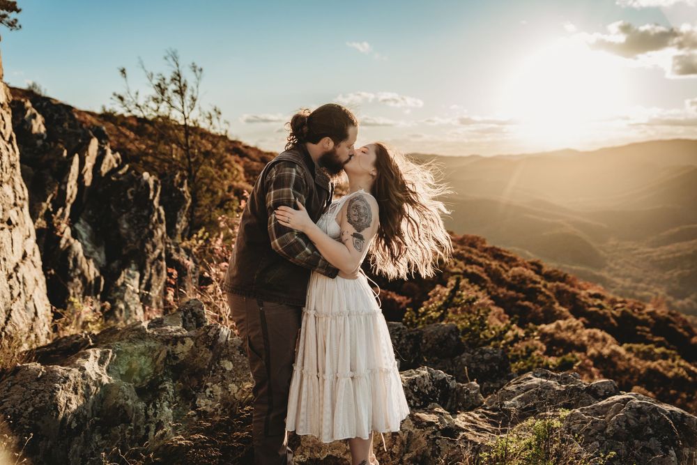 A couple embraces during sunset on a mountain cliff overlooking a scenic valley landscape.