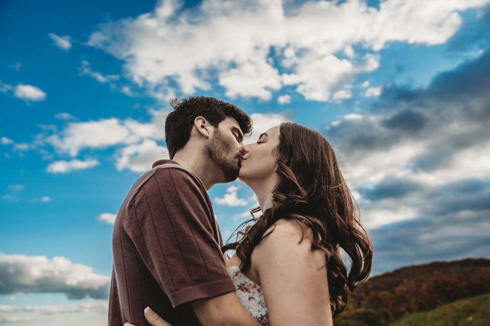 A romantic couple kisses outdoors against a dramatic cloudy sky during golden hour.