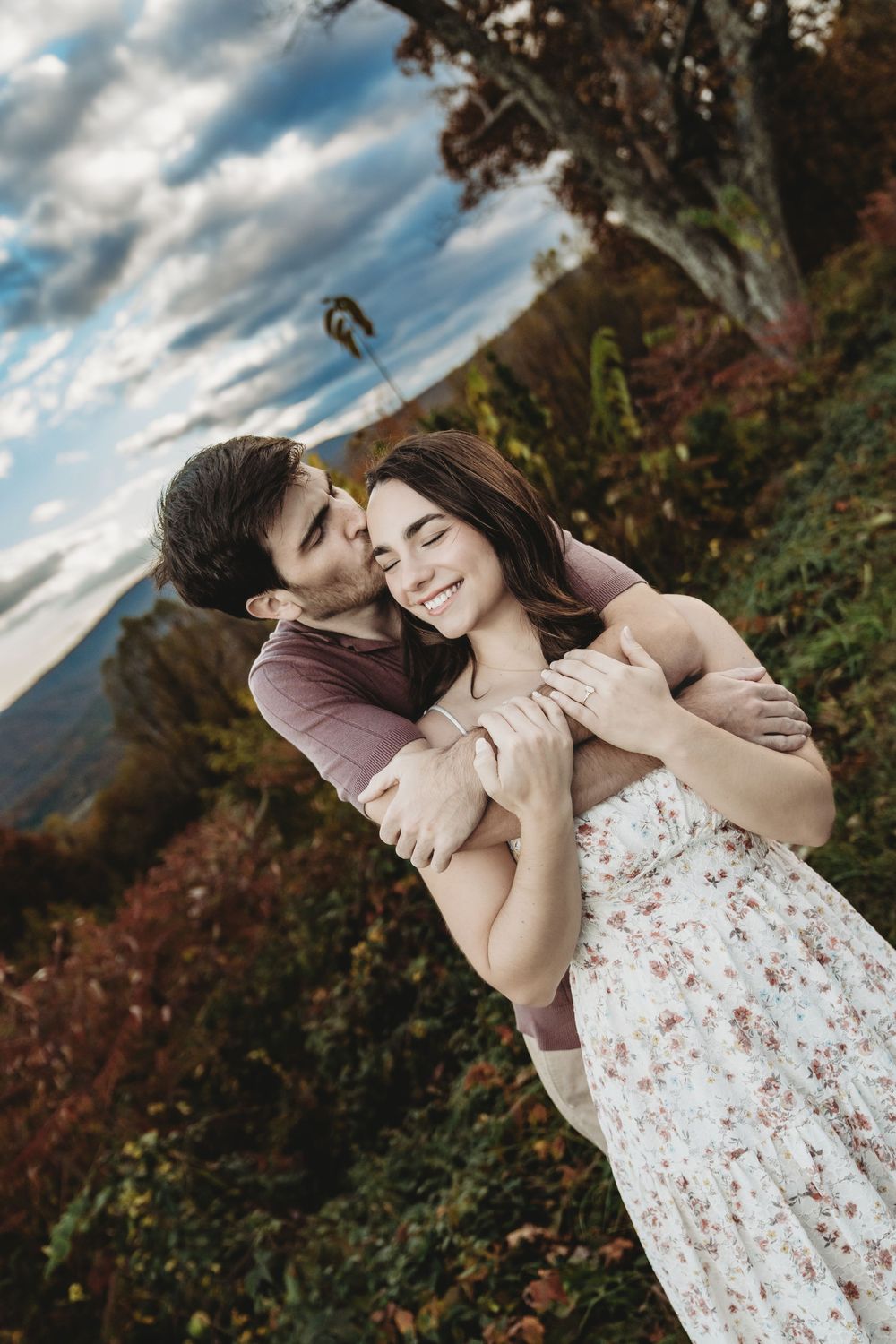 A romantic couple embraces against a scenic autumn mountain backdrop with dramatic clouds in the sky.