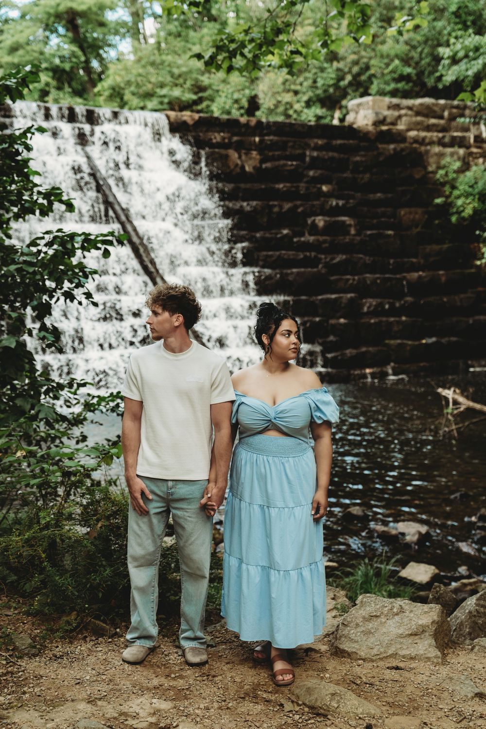 A couple stands together in front of a cascading waterfall surrounded by lush greenery and stone walls.