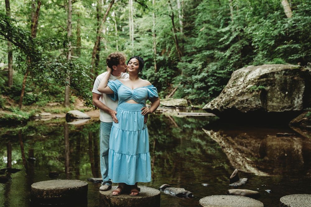 A group in blue dresses stands near a serene creek in a lush forest setting.