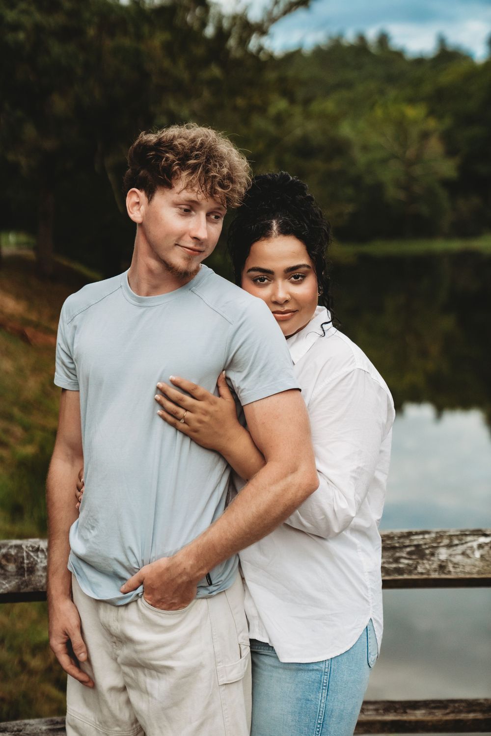 A romantic couple embraces by a serene lake on a sunny day, wearing casual white and blue outfits.