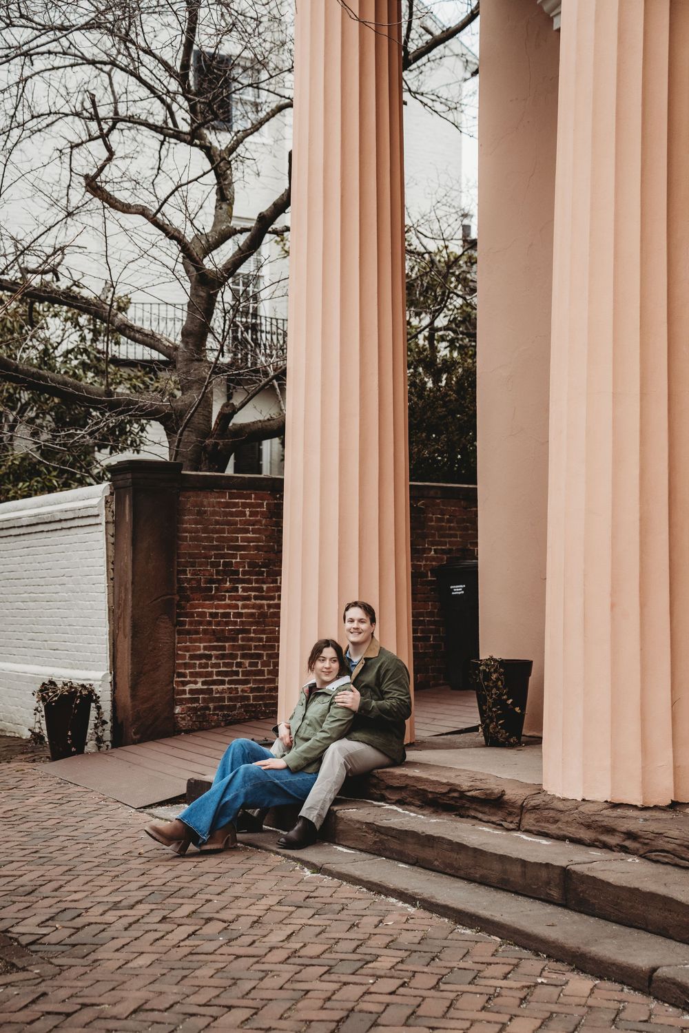 Couple sitting on brick steps beside large pink columns of a historic building surrounded by bare trees.