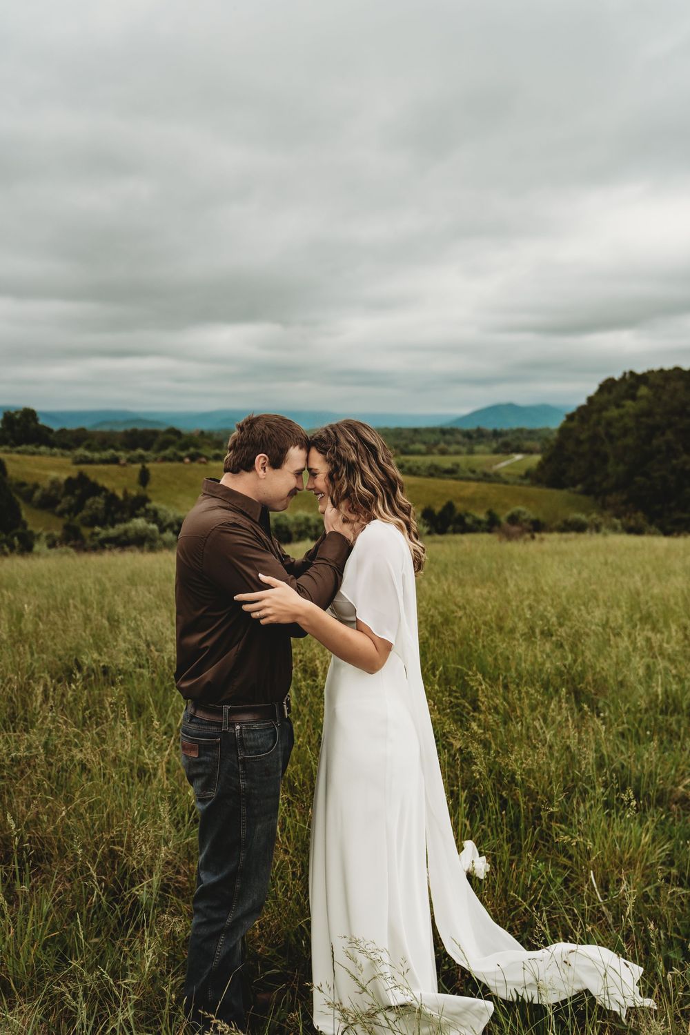 Couple in wedding attire sharing a romantic moment in a lush green field with mountain views under cloudy skies.
