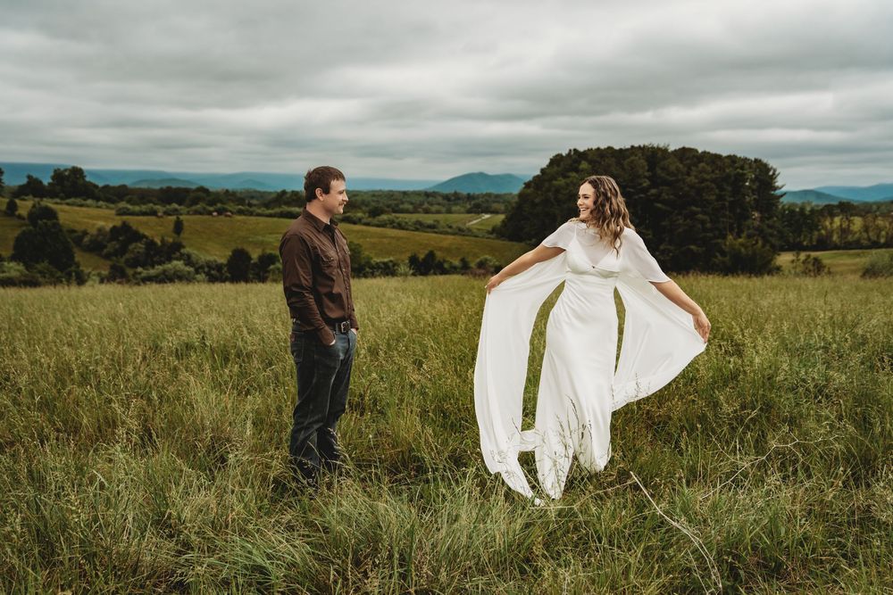 Couple in green field, bride in flowing white dress, groom in dark shirt, mountains backdrop, cloudy sky.