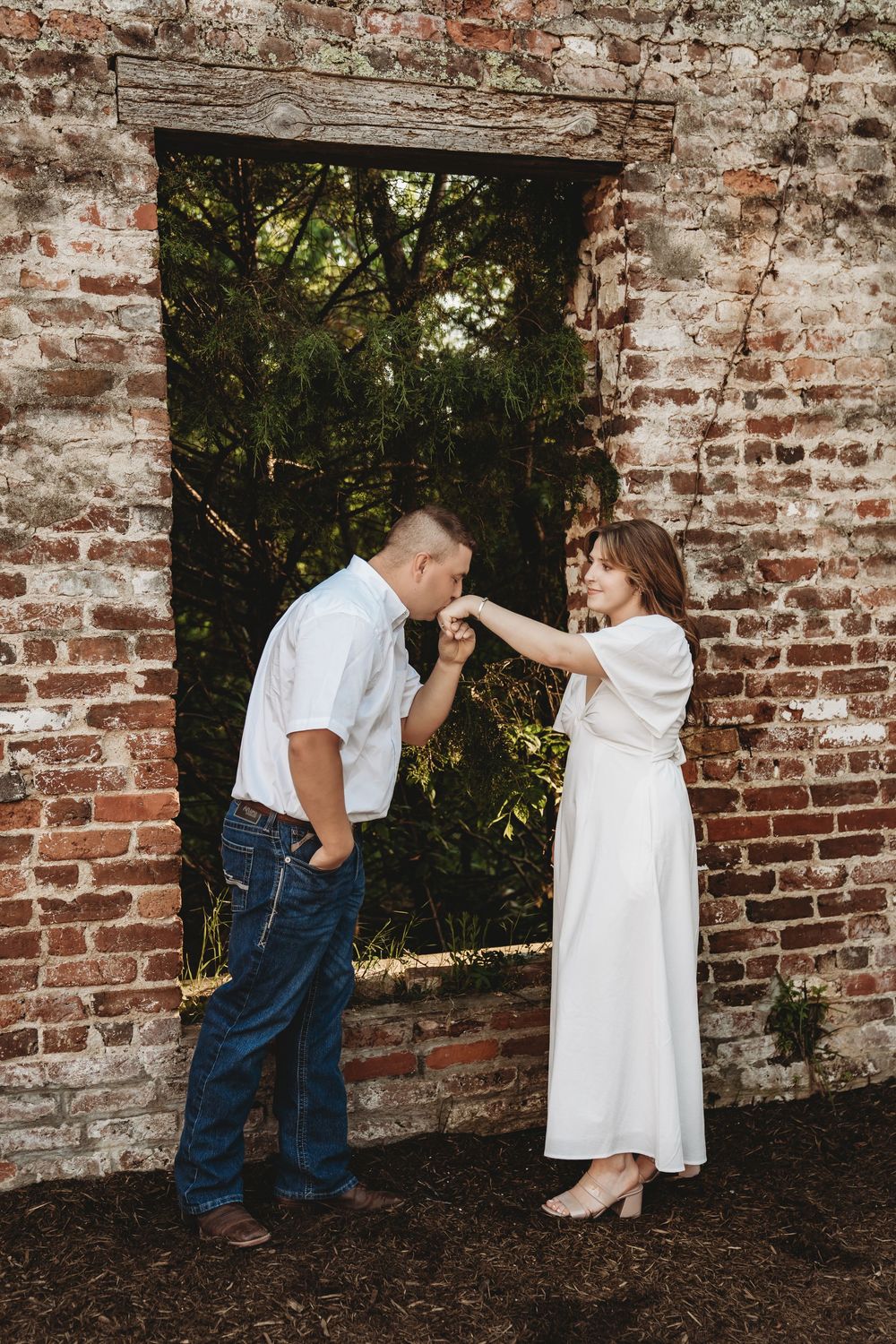 Man in white shirt kisses woman's hand in white dress by rustic brick wall doorway in romantic outdoor engagement photo.