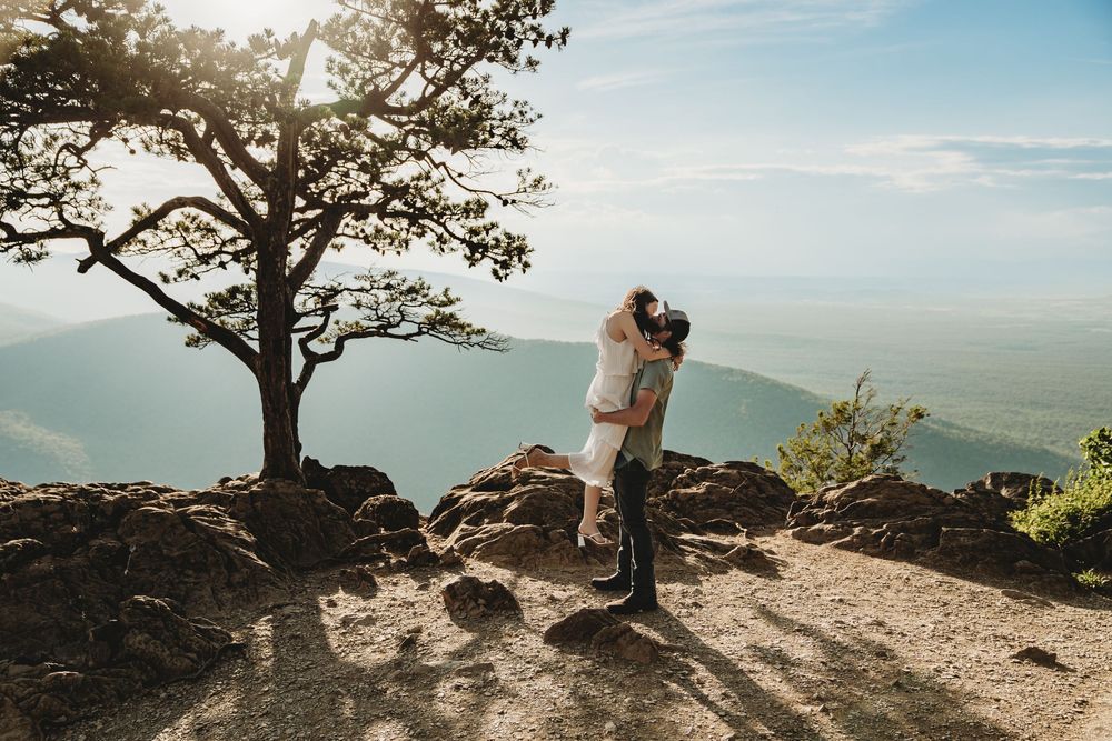 Couple sharing a romantic kiss on a rocky mountain overlook with scenic blue ridge valley views and sunlit tree backdrop.