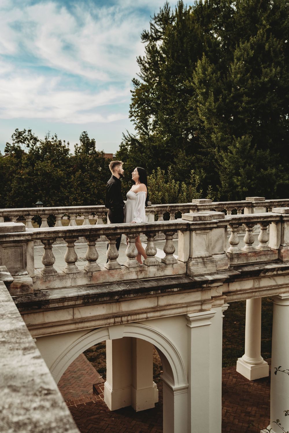 A couple stands on an elegant white stone balcony with classical architectural details surrounded by lush trees.