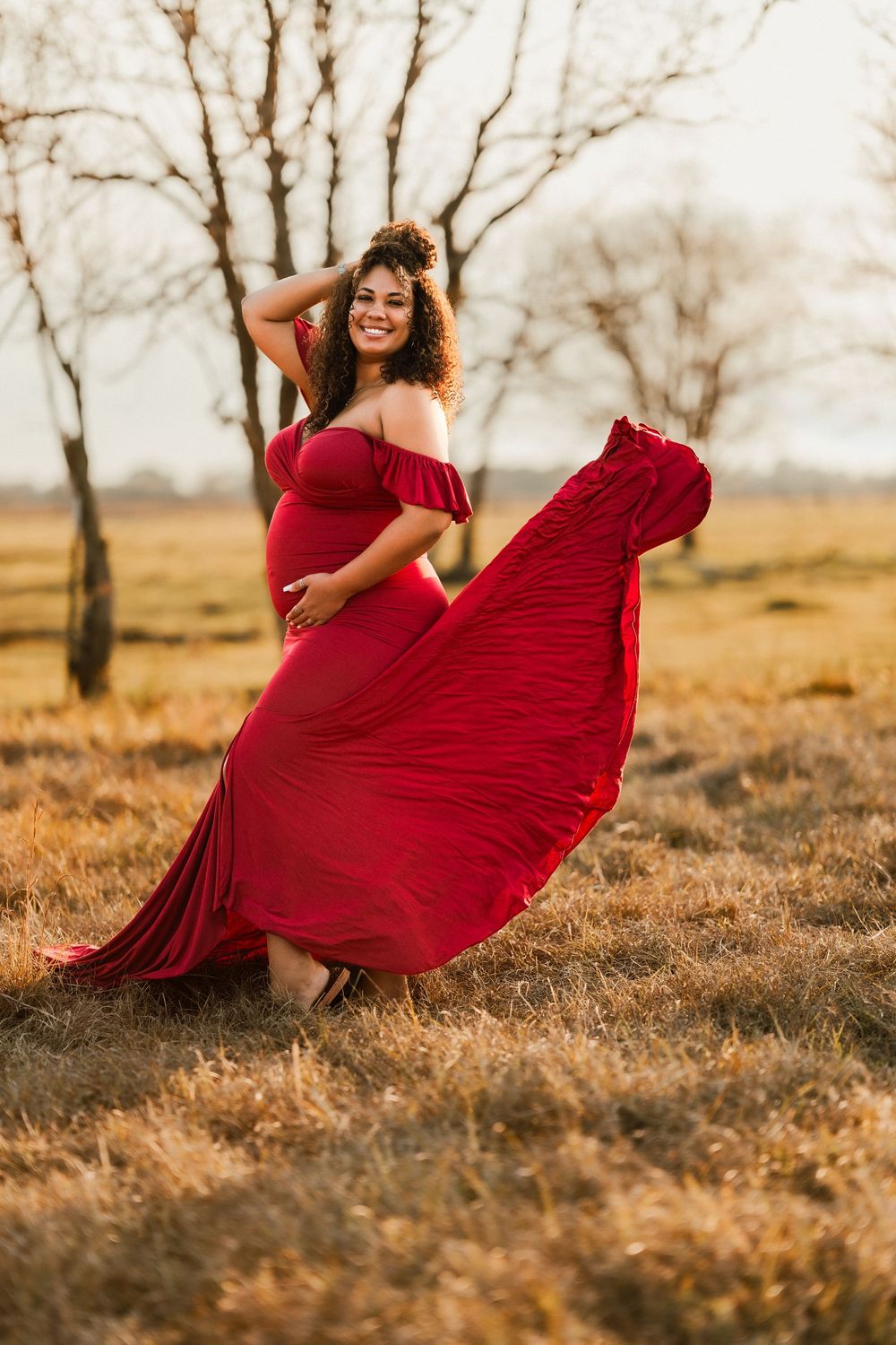 A flowing red dress twirls gracefully in golden sunlight against a backdrop of bare trees in an autumn field.
