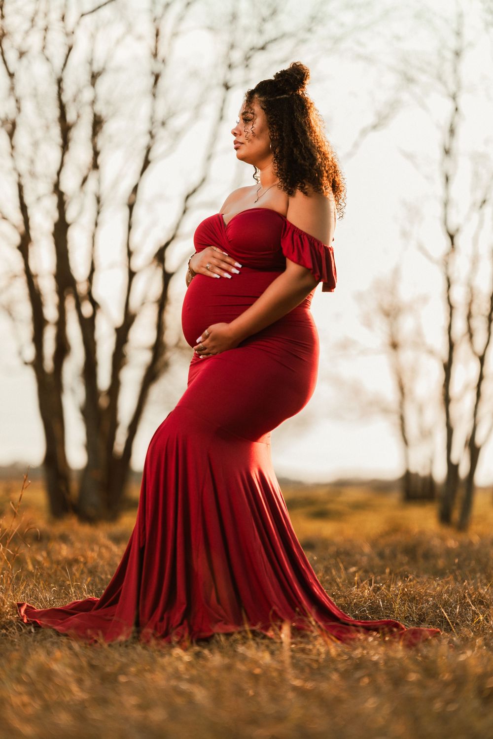 A pregnant woman in a flowing burgundy dress poses gracefully in an autumn forest setting during golden hour.