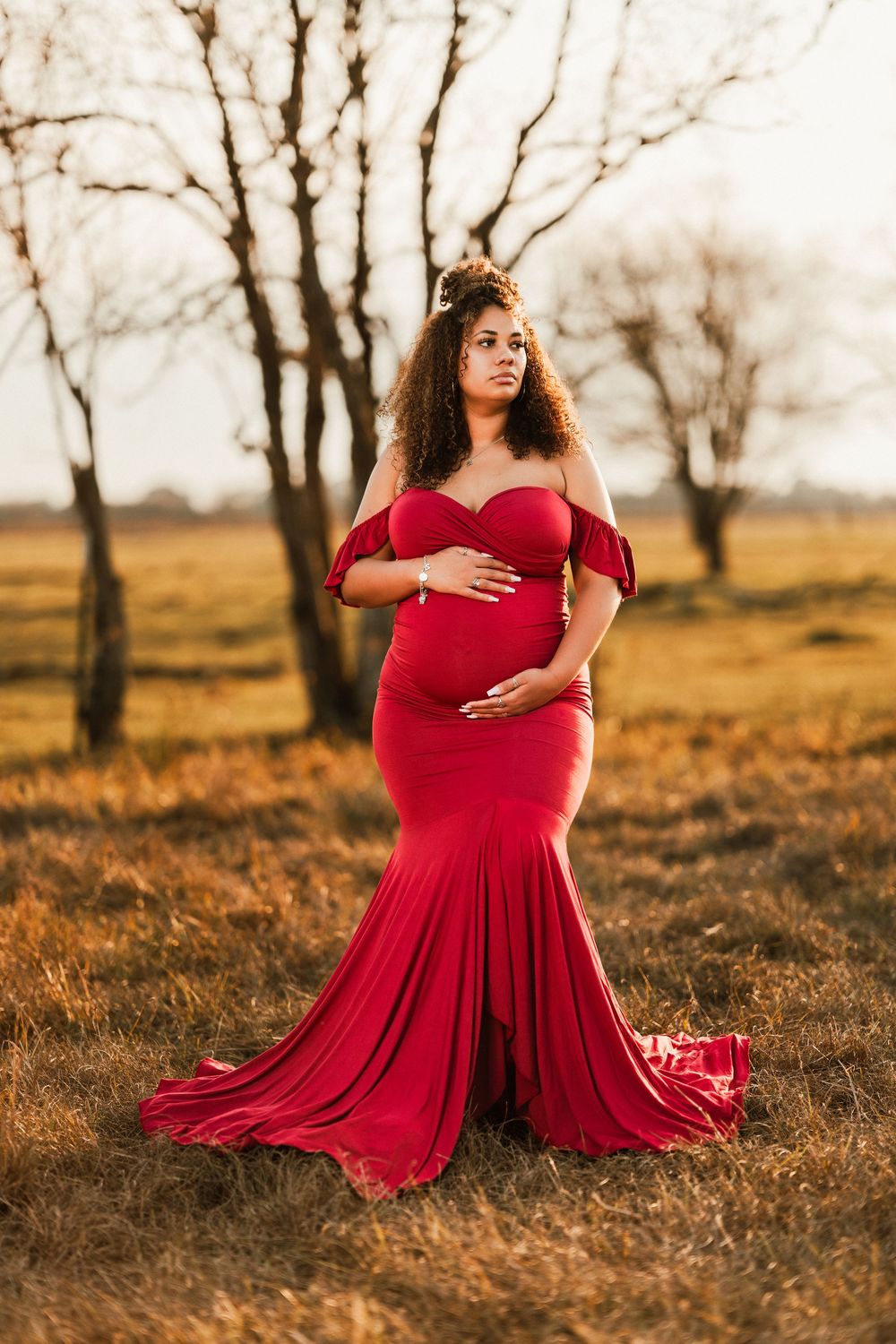 A woman in a flowing red dress poses gracefully in a golden autumn field with bare trees in the background.