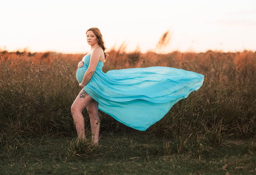 A pregnant woman in a flowing turquoise dress poses in a field at sunset as fabric billows dramatically in the wind.